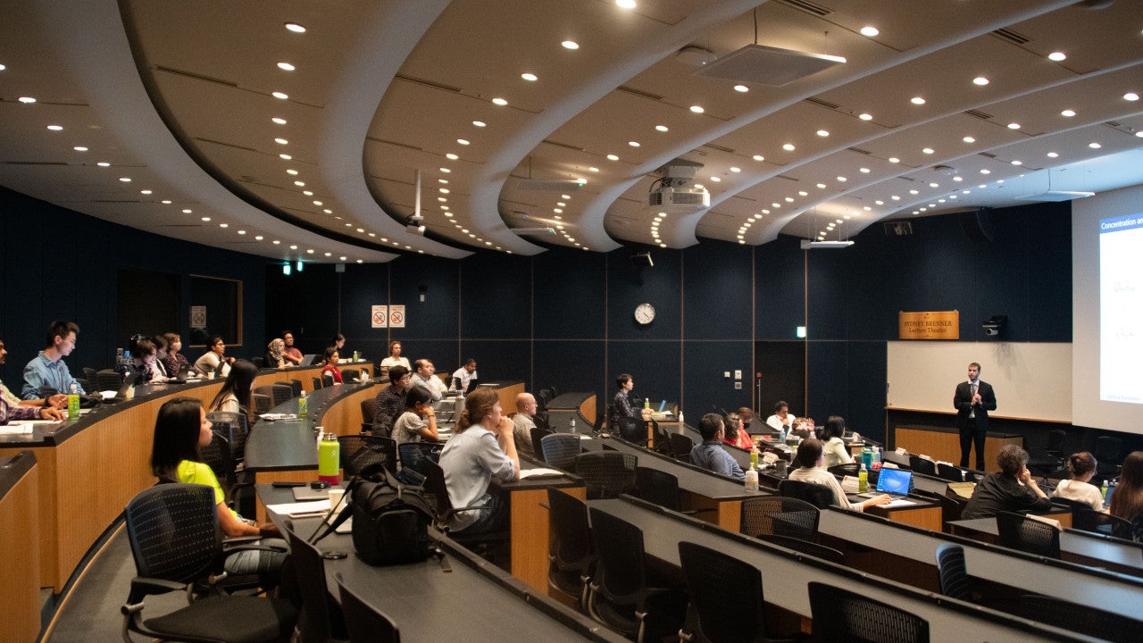 Numerous attendees sitting in chairs at desks in a seminar hall. One speaker is standing in front of the screen at the right.