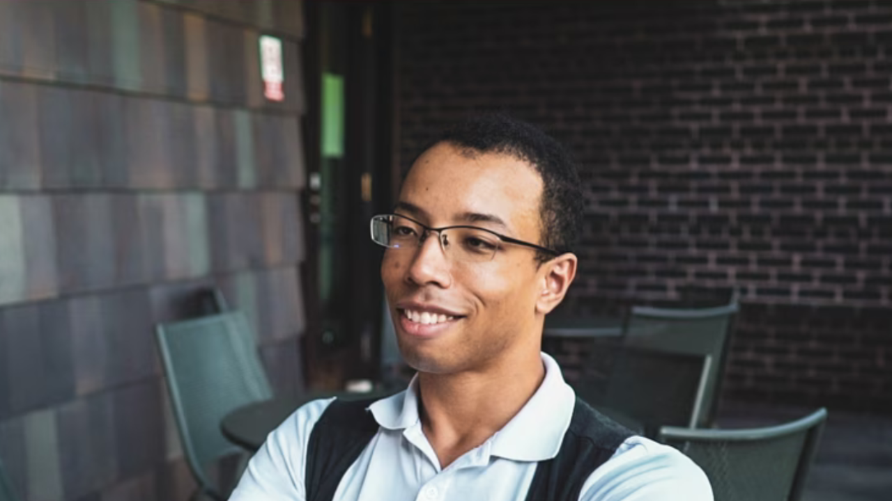 Man with glasses and blue shirt sitting at table with brick wall behind him