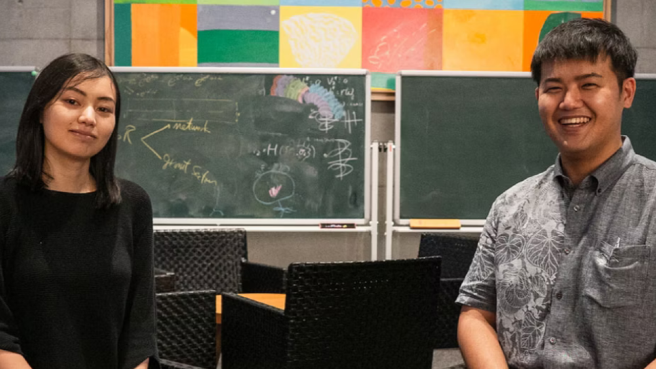 Two students sitting on chairs with chalkboard and black chairs in background