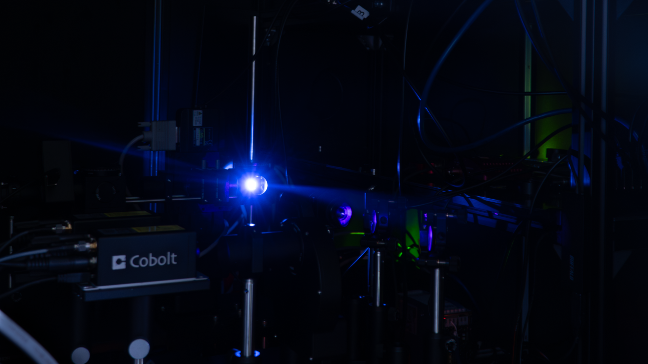 A photograph of a blue laser light hitting a reflective surface inside a complicated spectroscopy setup, housed in a dark area.
