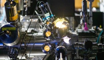Photograph of a section of a large, silvery machine, with circular openings like portholes emitting bright lights. Multiple tubes and wires are attached, and more machinery is visible in the background.