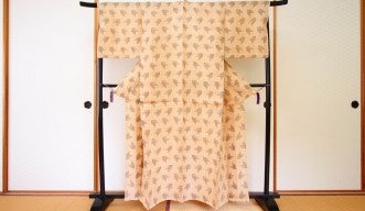 Photograph of a tan kimono suspended on a dark, wooden rack standing on tatami mats, in front of fusuma room dividers.