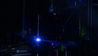 A photograph of a blue laser light hitting a reflective surface inside a complicated spectroscopy setup, housed in a dark area. 