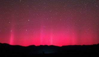 澄み切った夜空を背景に、山々のシルエットの上に広がる赤いオーロラの写真。