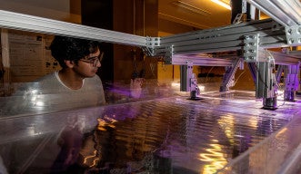 A scientist stands next to a giant water tank and looks at waves on the water’s surface.