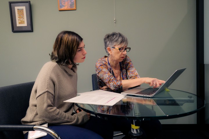 A student (left) and professor (right) looking at something on a laptop while they sit together at a glass table.