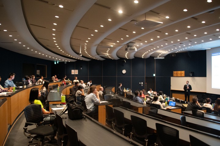 Numerous attendees sitting in chairs at desks in a seminar hall. One speaker is standing in front of the screen at the right.