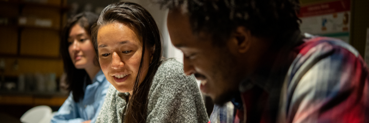 Three students sit next to each other, smiling, looking down to something off screen