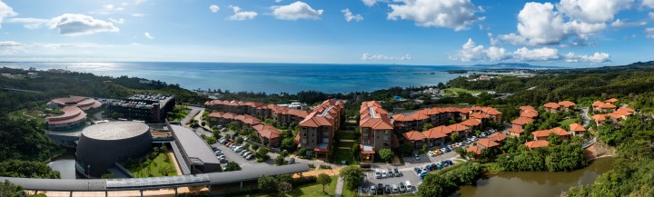 Aerial (drone) view of OIST's Village Center and entrance to the Tunnel Gallery, overlooking the ocean and a sunny sky with few clouds