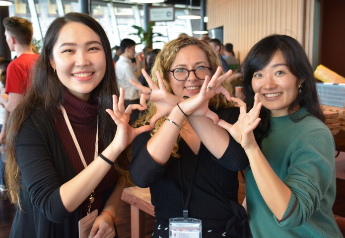 3 people in front posing with hands in shapes of hearts. People aslo in background.