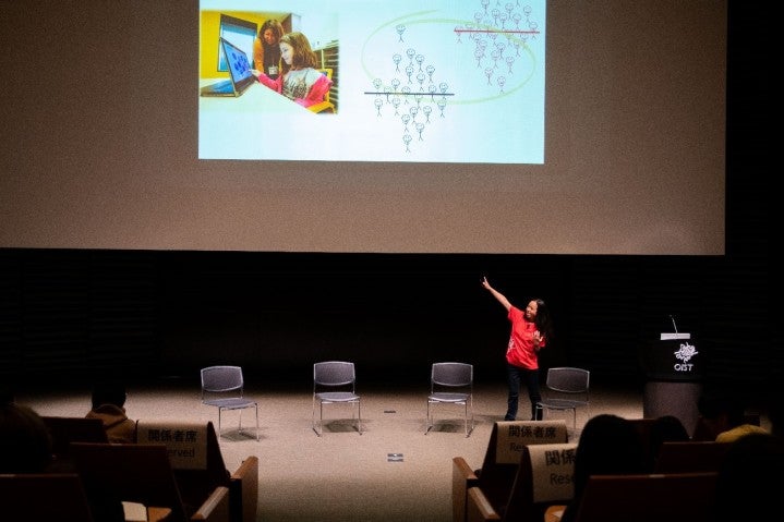 Researcher in a red OIST shirt pointing at the large screen, which shows the effect of social exclusion for people with ADHD.
