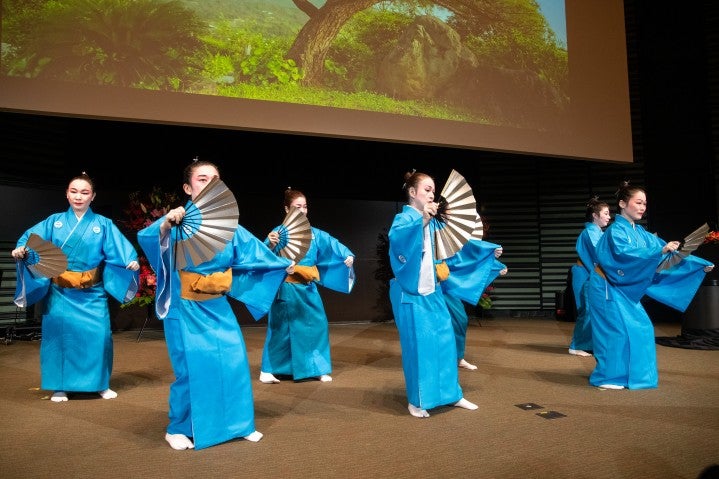 鮮やかな青の衣装をまとった舞踊家が、扇を広げて伝統的な沖縄の舞踊（琉球舞踊）を踊っている。