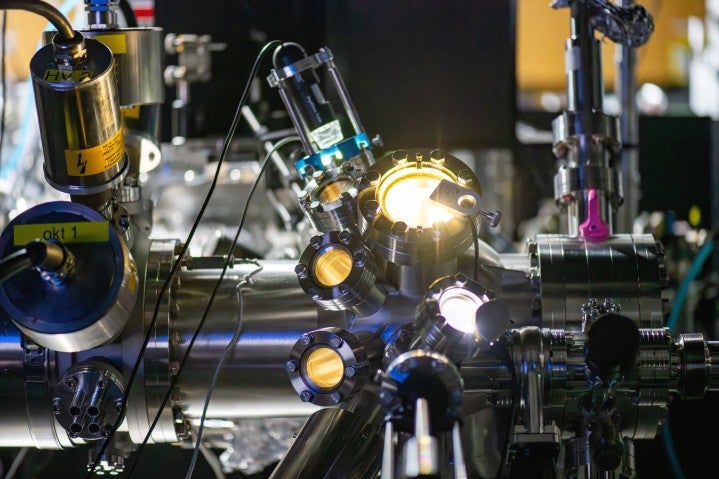 銀色の大型機械の円形開口部から明るい光が放出されている様子を写した写真。多数のチューブや配線が取り付けられており、背景にも多くの機器が並んでいる。