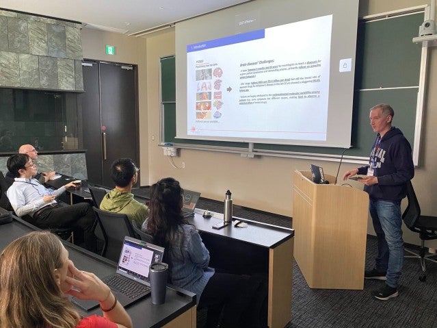 A speaker presenting in front of a screen to an audience seated in a lecture room