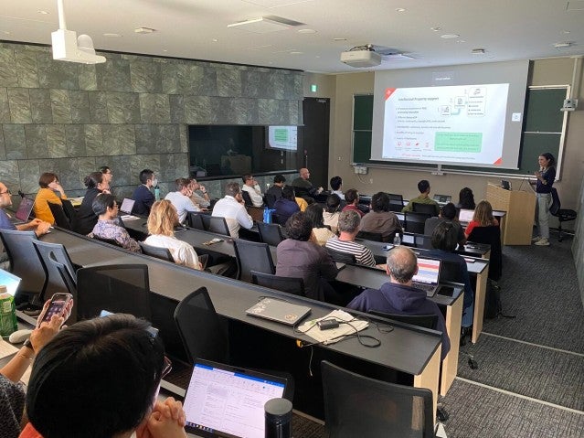 A speaker presenting in front of a screen to an audience seated in a lecture room