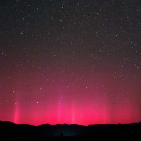 澄み切った夜空を背景に、山々のシルエットの上に広がる赤いオーロラの写真。