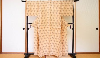 Photograph of a tan kimono suspended on a dark, wooden rack standing on tatami mats, in front of fusuma room dividers.