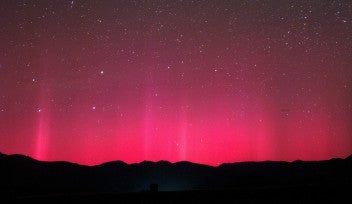 澄み切った夜空を背景に、山々のシルエットの上に広がる赤いオーロラの写真。