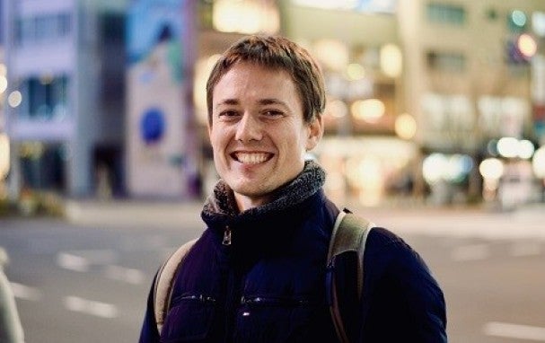 Man wearing backpack and thick jacket. Outdoors near street, smiling