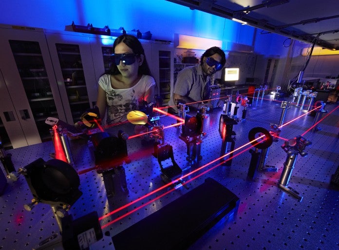two students wearing dark sunglasses in blue lighting lab. Red lasers bouncing around on mirrors in front of them.