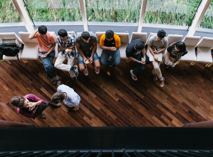 View looking down on floor below in cafe. Seven people sitting in chairs and two standing chatting. Most drinking from cups.