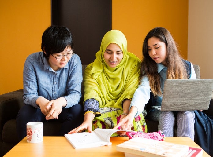 three people sitting on a sofa. Right one has a laptop on their lap and is pointing to a book on a desk in front of them.