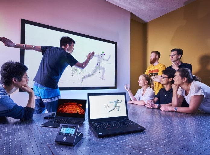 5 people standing and sitting on the right looking at screen in center. Screen has person's model in running motion. Two people on left, one doing running motion. Two laptops in foreground.